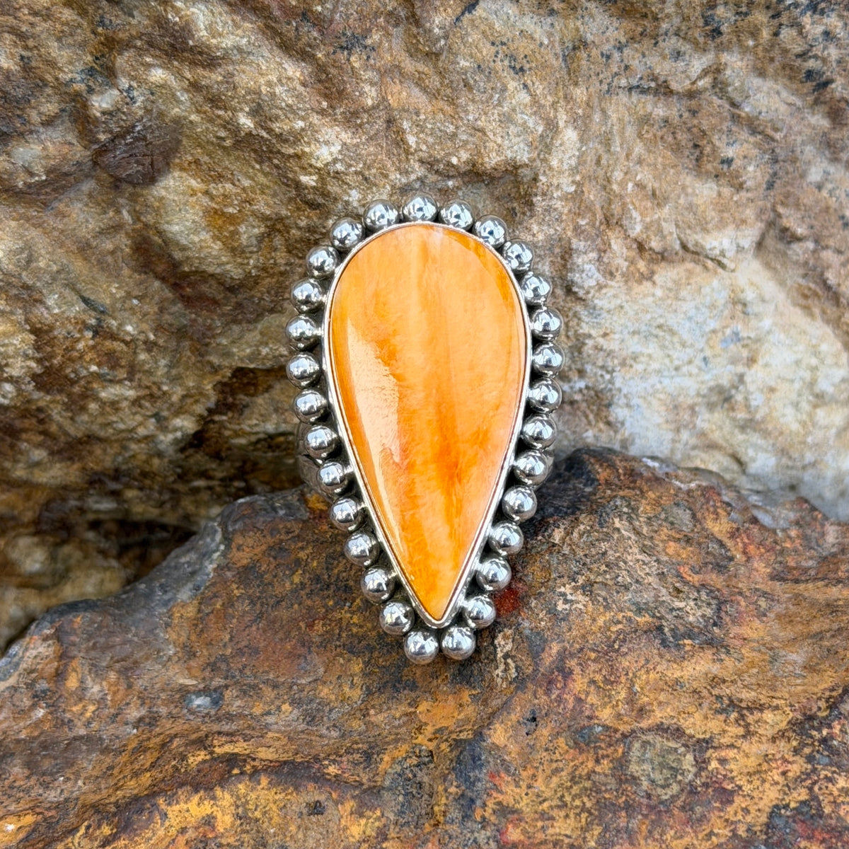 A sterling silver ring by Artie  Yellowhorse that holds an upside down teardrop cabochon made of orange spiny oyster. It is surrounded by silver beads. It rests on a rocky surface