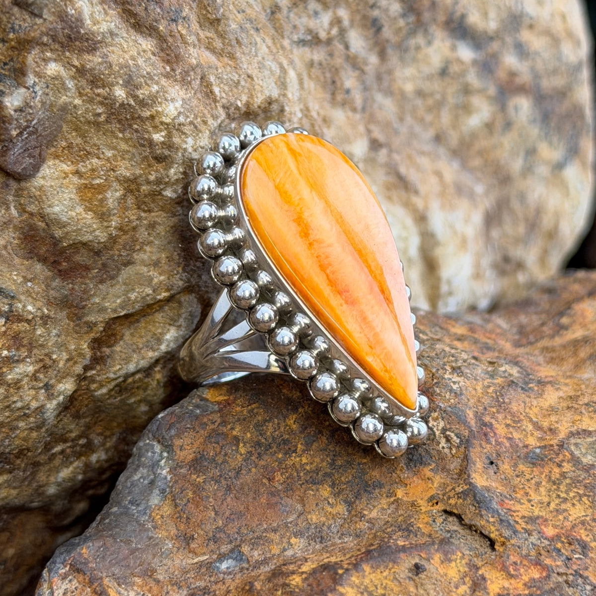 A sterling silver ring by Artie  Yellowhorse that holds an upside down teardrop cabochon made of orange spiny oyster. It is surrounded by silver beads. It rests on a rocky surface