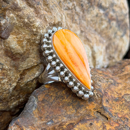 A sterling silver ring by Artie  Yellowhorse that holds an upside down teardrop cabochon made of orange spiny oyster. It is surrounded by silver beads. It rests on a rocky surface