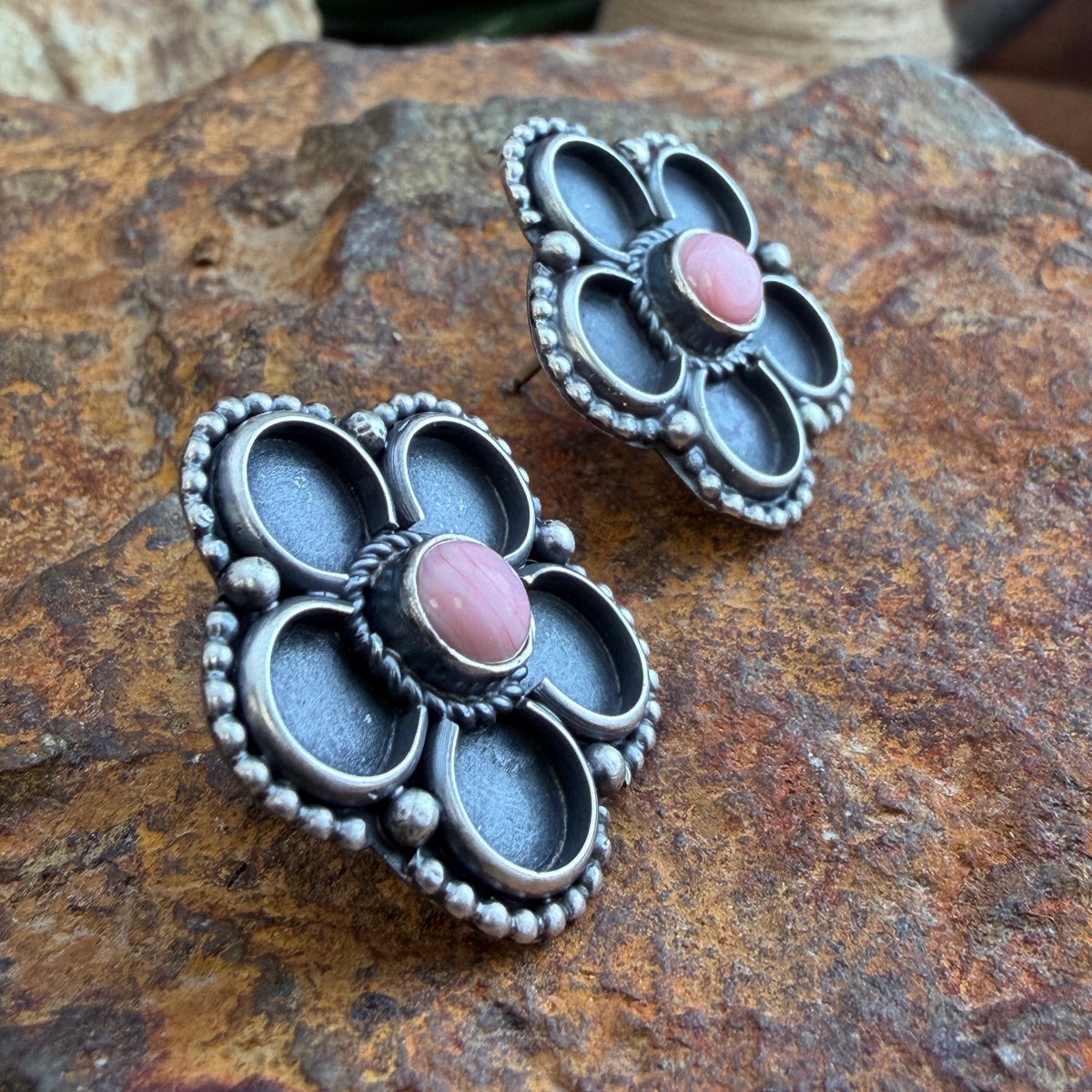 A pair of silver-toned flower-shaped earrings with oval pink cabochon stones, displayed on a textured brown rock surface.