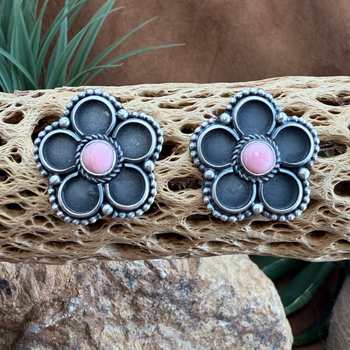 A pair of silver-toned flower-shaped earrings with dark gray petal inlays and pink cabochon centers, displayed on a piece of driftwood with rocks and greenery in the background.