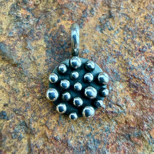 A textured sterling silver pendant with a cluster of raised metallic beads, resting on a rustic, textured brown and gold stone surface.