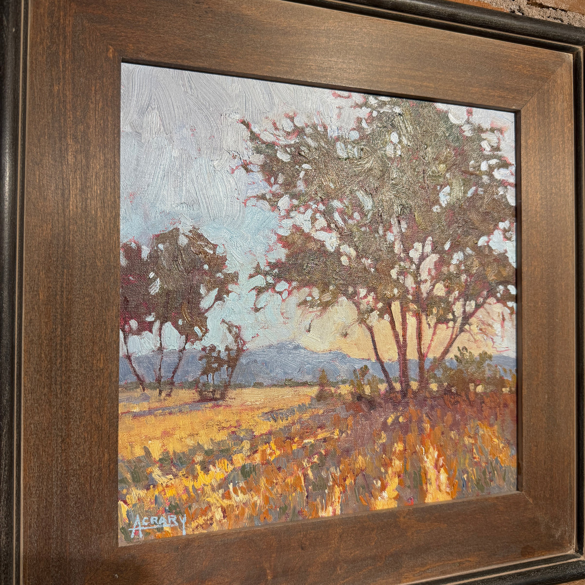 A framed landscape oil painting featuring trees, a field, and distant hills, housed in a dark brown wooden frame.