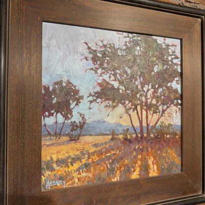 A framed landscape oil painting featuring trees, a field, and distant hills, housed in a dark brown wooden frame.