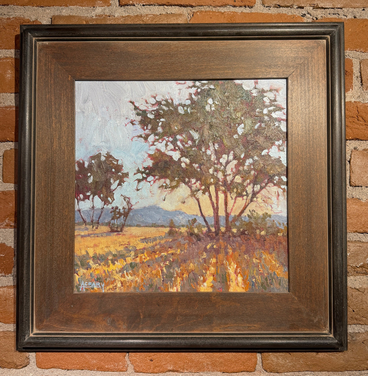 A framed landscape oil painting featuring trees and a field, mounted on a rustic red brick wall. The painting is housed in a dark brown wooden frame.