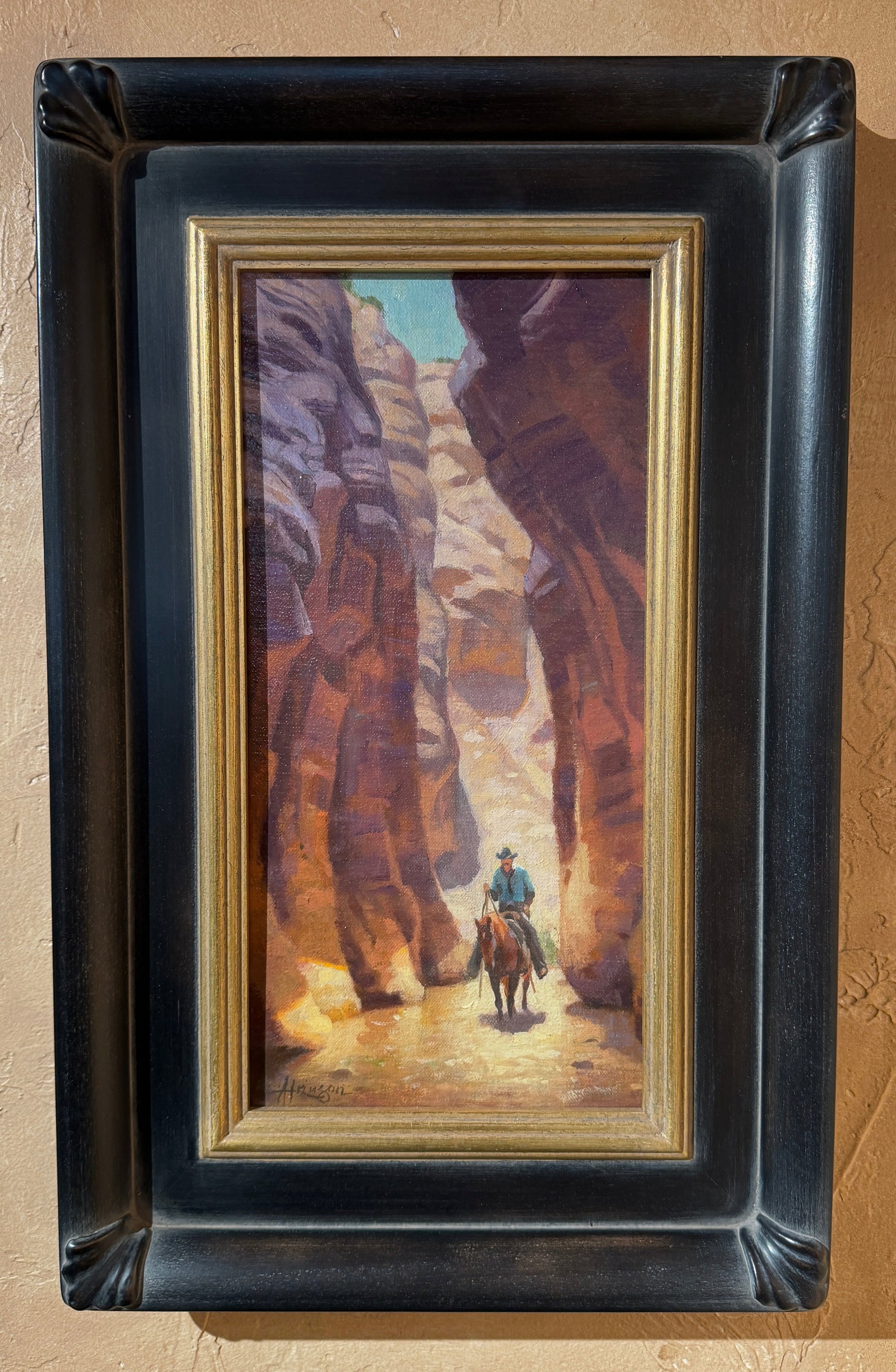 A framed landscape painting of a cowboy riding through a canyon, mounted on a textured beige wall. The painting features warm brown and orange canyon walls with a small figure of the cowboy in blue and green.