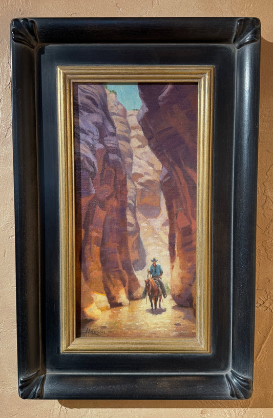 A framed landscape painting of a cowboy riding through a canyon, mounted on a textured beige wall. The painting features warm brown and orange canyon walls with a small figure of the cowboy in blue and green.