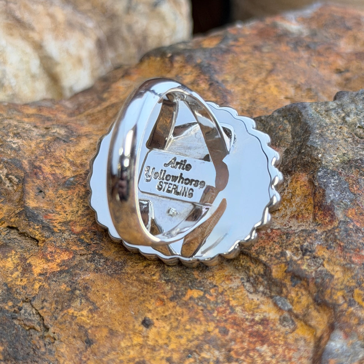 Sterling Silver ring by Artie Yellowhorse. large oval dark blue cabochon surrounded by rope border and silver beads. it rests on a rocky surface. this one showcases the back of the ring with stamped artist and "sterling"