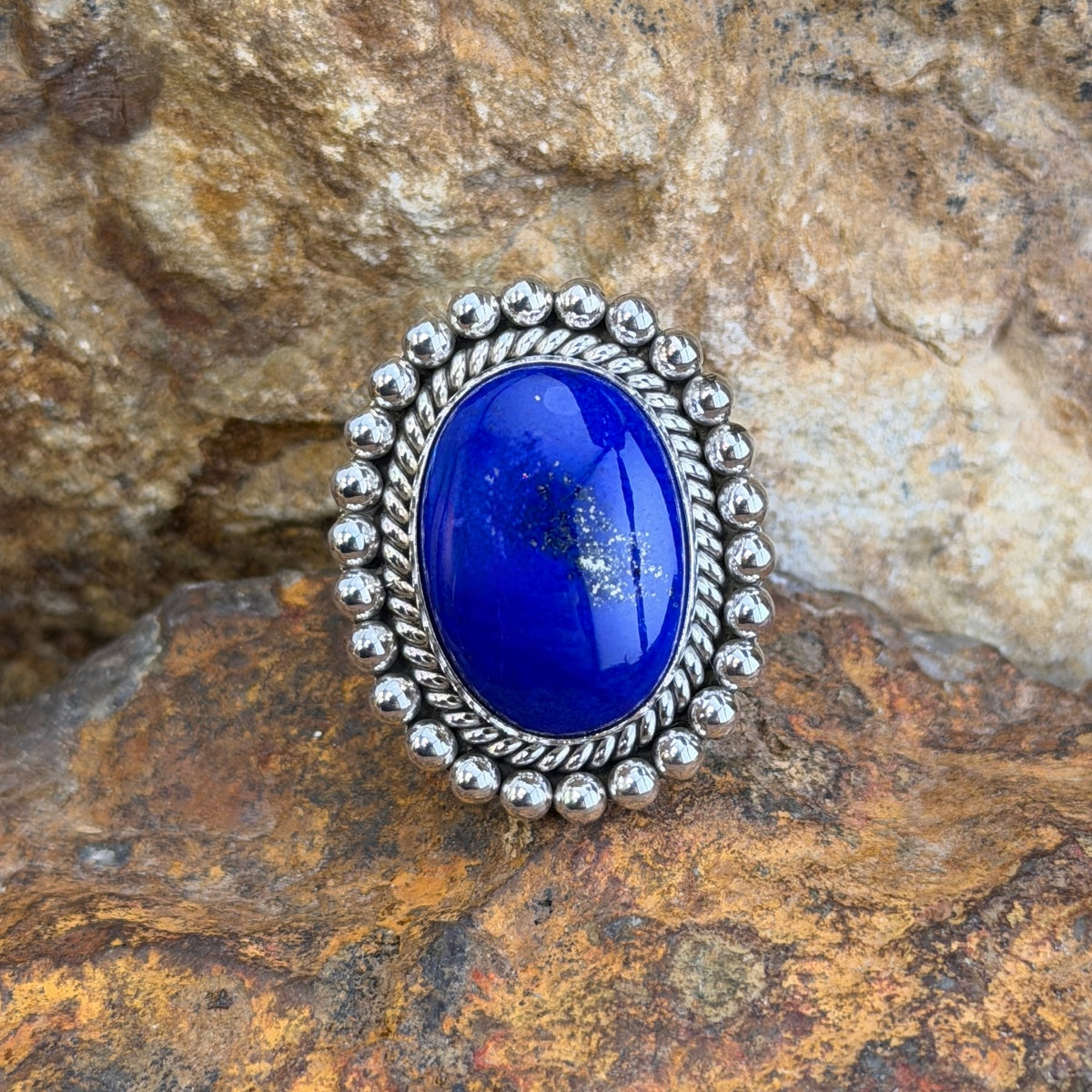 Sterling Silver ring by Artie Yellowhorse. large oval dark blue cabochon surrounded by rope border and silver beads. it rests on a rocky surface