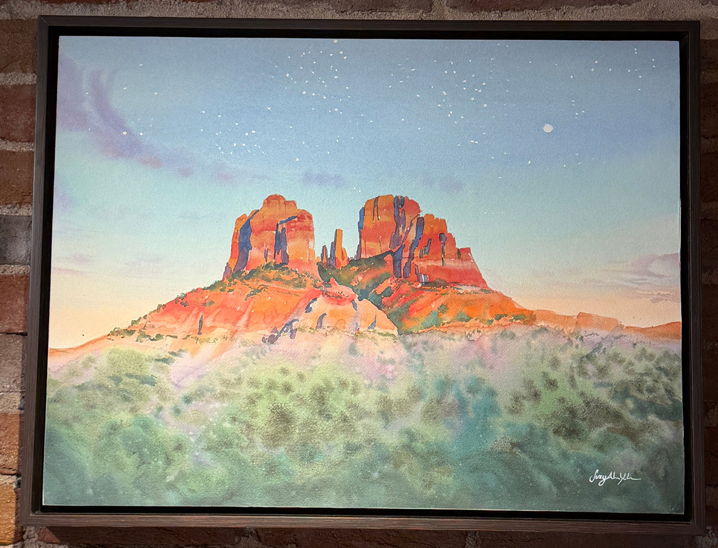 Framed painting of a desert landscape with red rock formations on a brick wall.