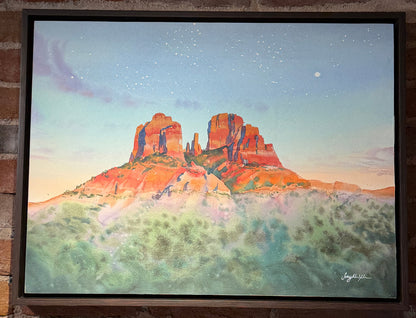 Framed painting of a desert landscape with red rock formations on a brick wall.