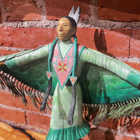 Decorative figure with a green garment and pink sash against a brick wall