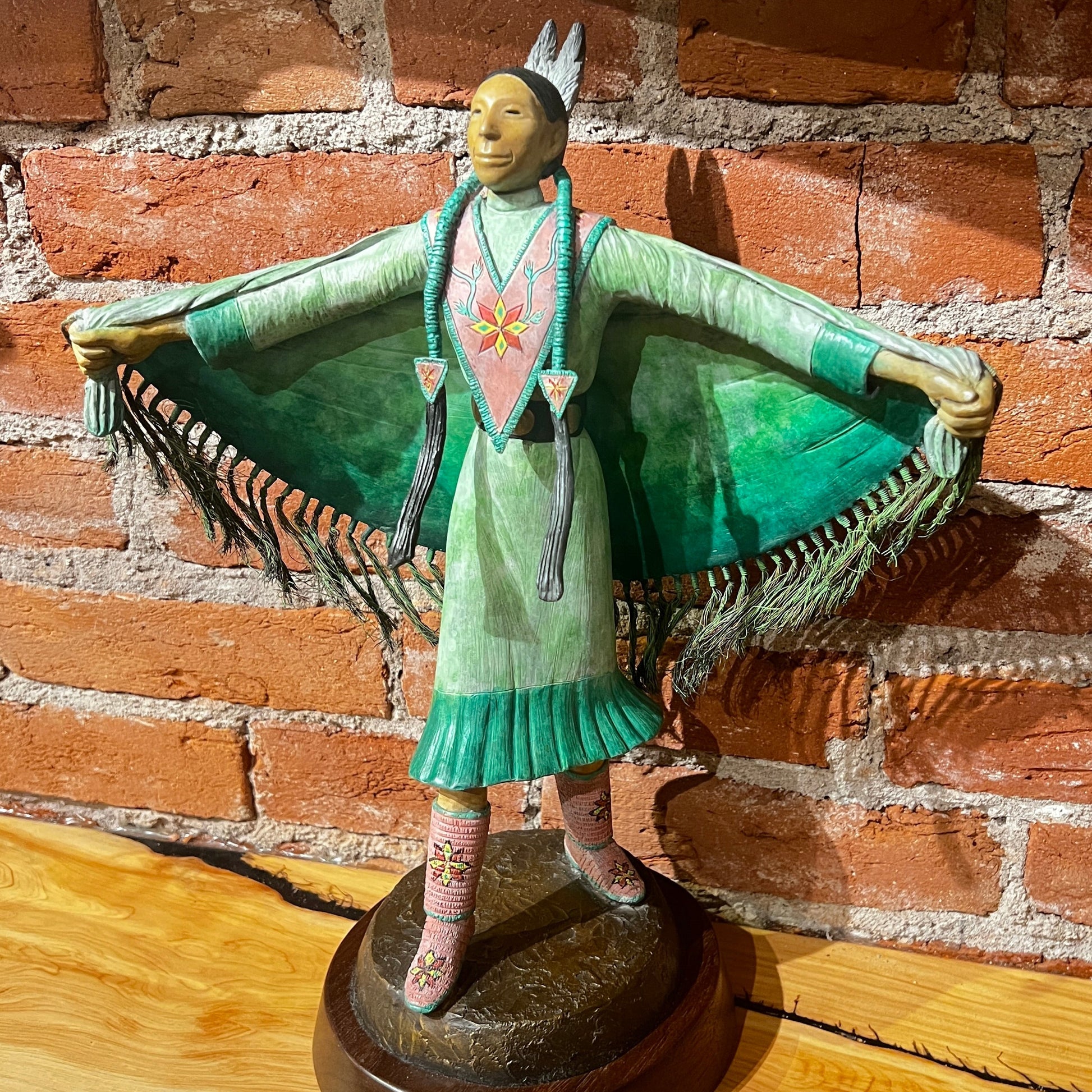 Decorative figurine of a person in a green dress with outstretched arms against a brick wall.