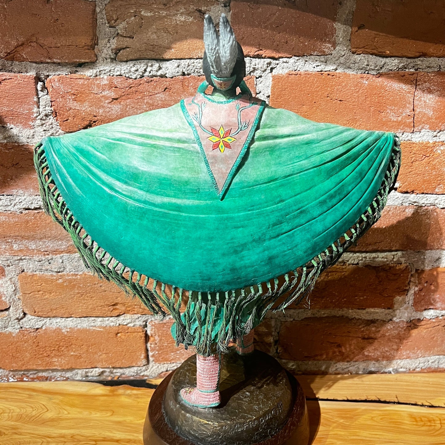 Decorative figure with outstretched arms wearing a green shawl with green against brick wall.