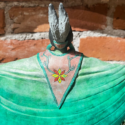 Statue of a figure with green garment and headdress against a brick wall.