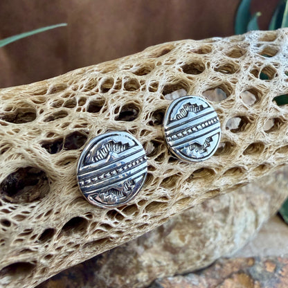 A pair of round silver-toned stud earrings with engraved geometric patterns, displayed on a textured natural cactus bone with rocks and greenery in the background.