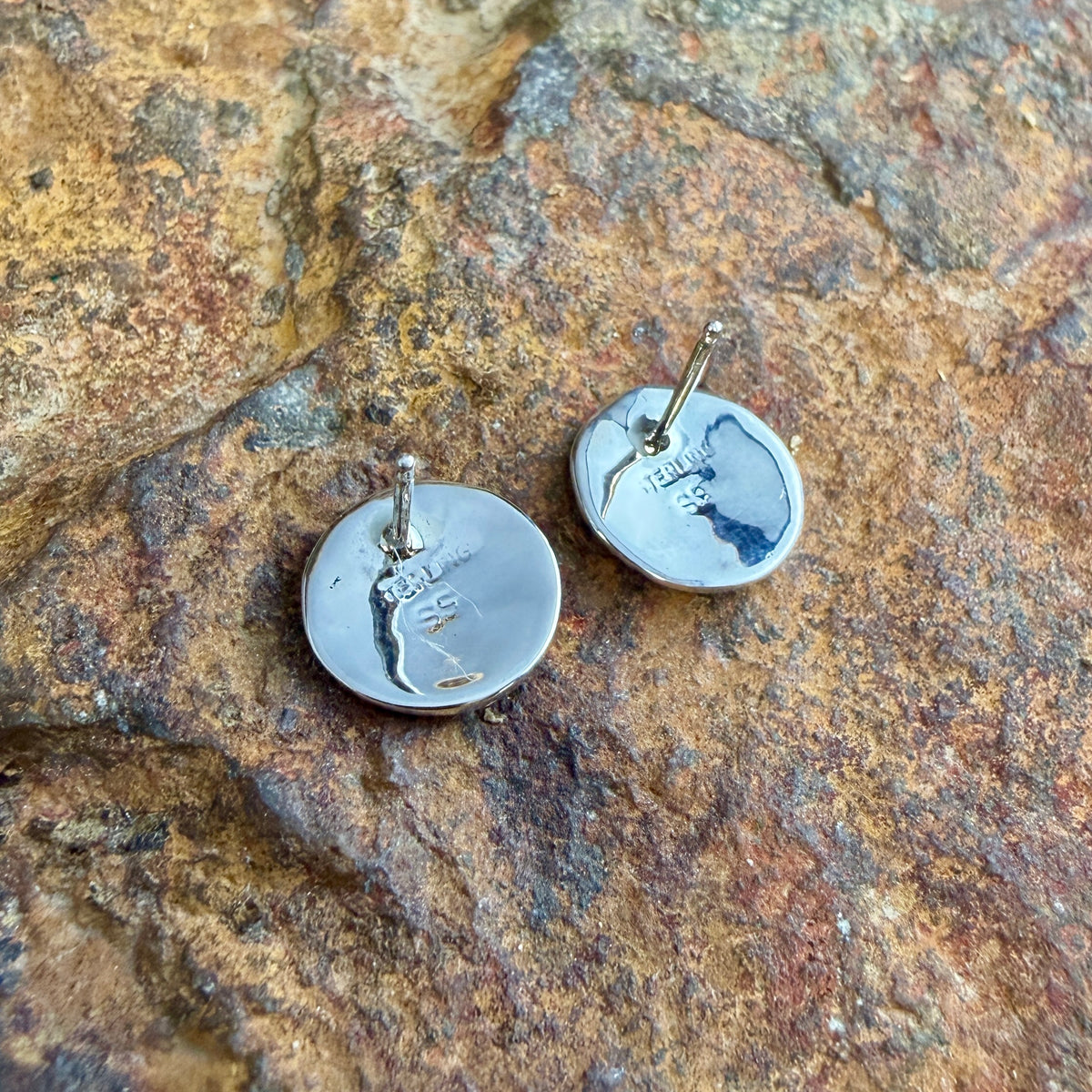 Two round silver disc earrings with post backs, photographed on a textured brown and gold rock surface. 