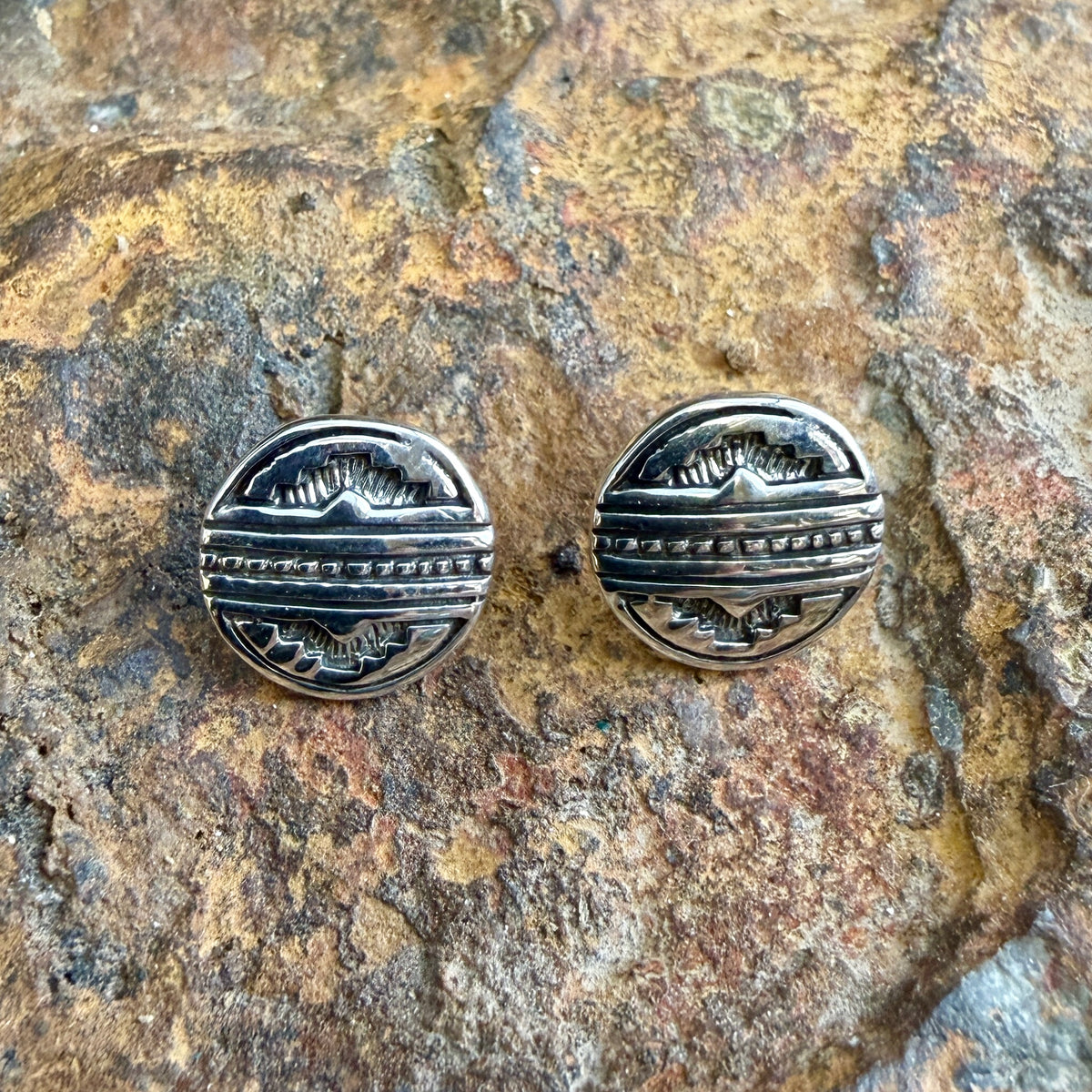 A pair of round sterling silver stud earrings with engraved geometric patterns, displayed on a textured brown and gold rock surface.
