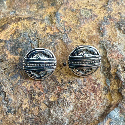 A pair of round sterling silver stud earrings with engraved geometric patterns, displayed on a textured brown and gold rock surface.