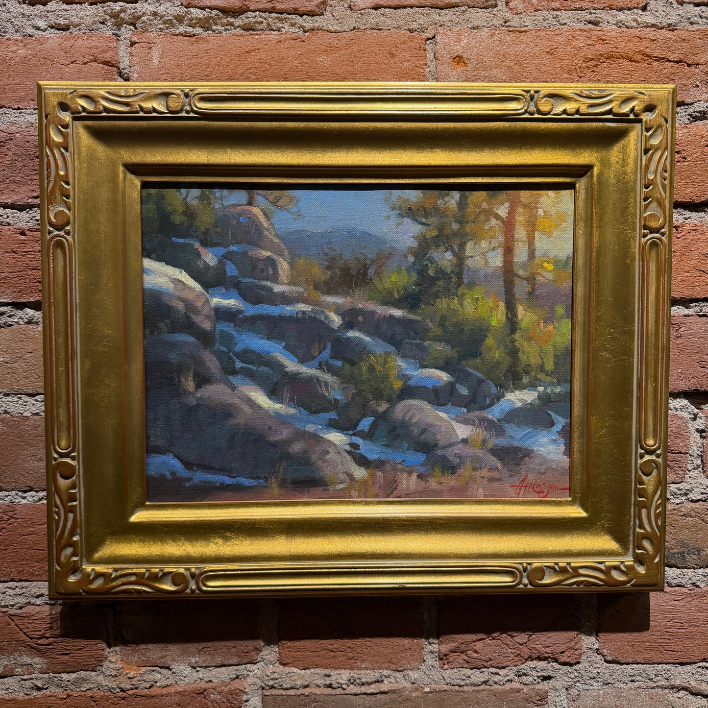 A framed landscape painting of a rocky stream with trees and distant mountains, set in an ornate gold frame and mounted on a red brick wall.