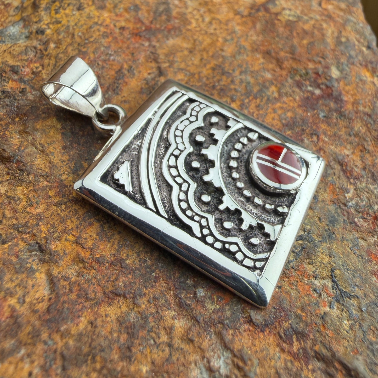 Silver pendant with intricate designs and a red stone on a textured brown surface