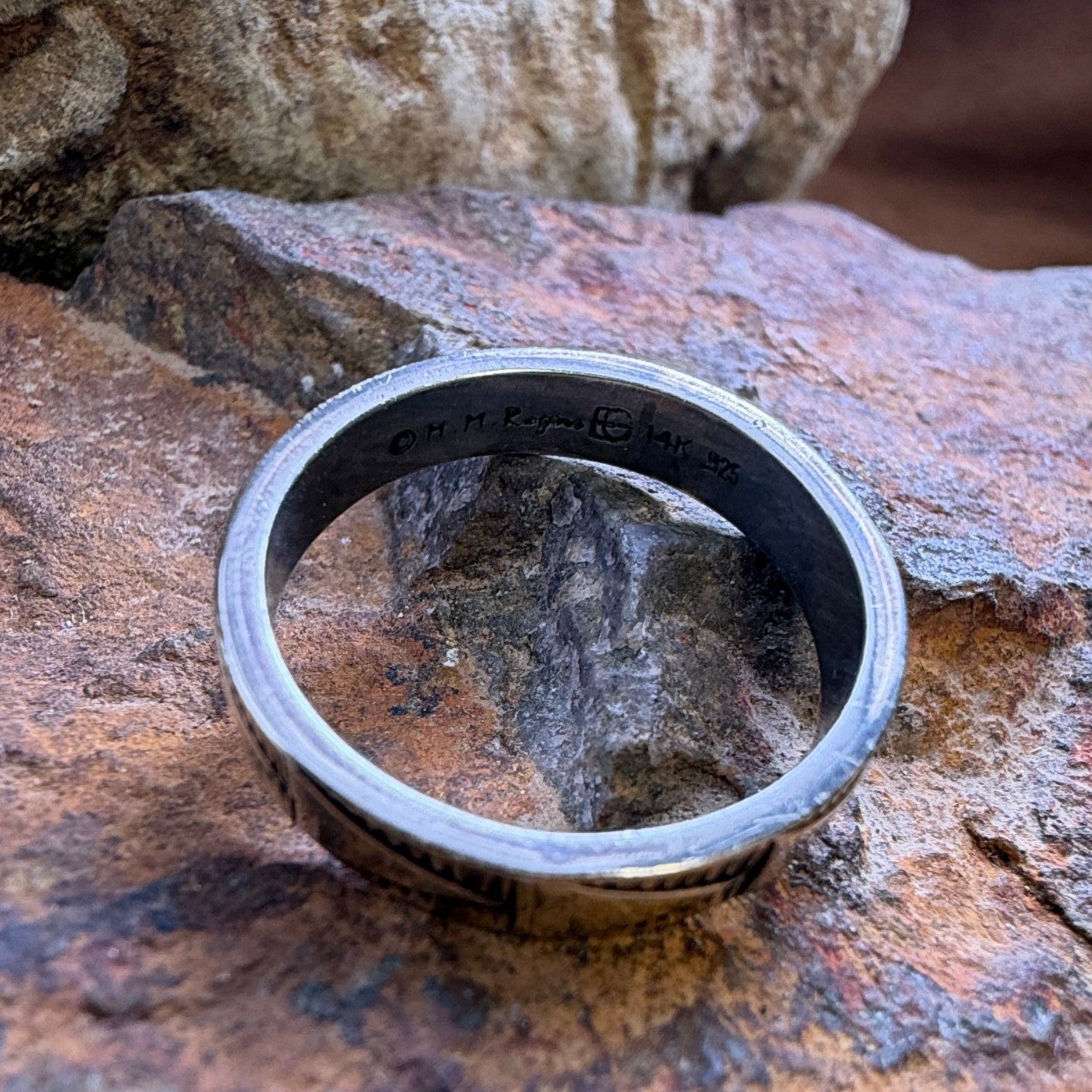 A silver-toned metal band ring with engraved text, resting on a textured brown and orange rock outdoors.
