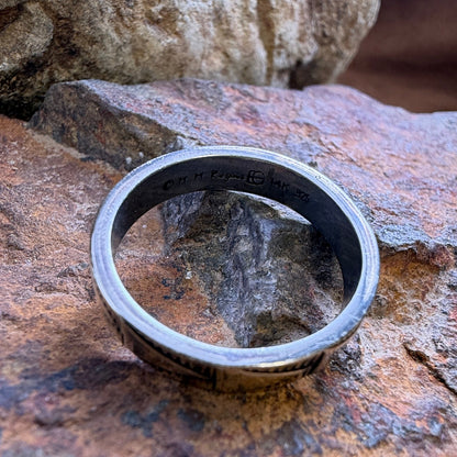 A silver-toned metal band ring with engraved text, resting on a textured brown and orange rock outdoors.
