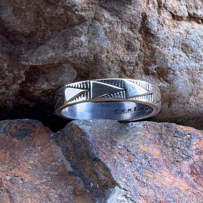 A silver band ring with geometric engraved patterns, resting on a textured orange-brown rock against a larger brown rock background.