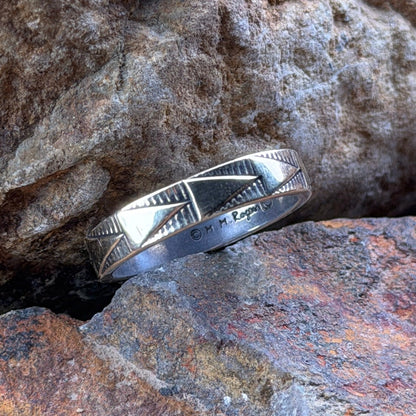 A silver band ring with a textured, geometric patterned interior, resting on textured brown and gray rocks.