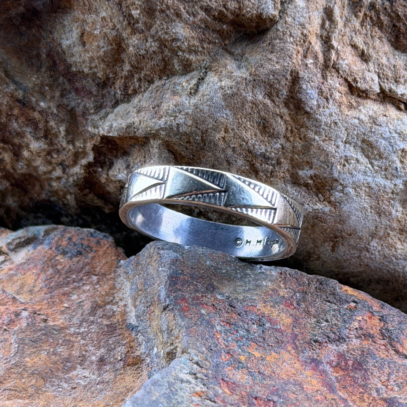 A silver band ring with engraved geometric patterns, resting on textured orange and brown rocks.