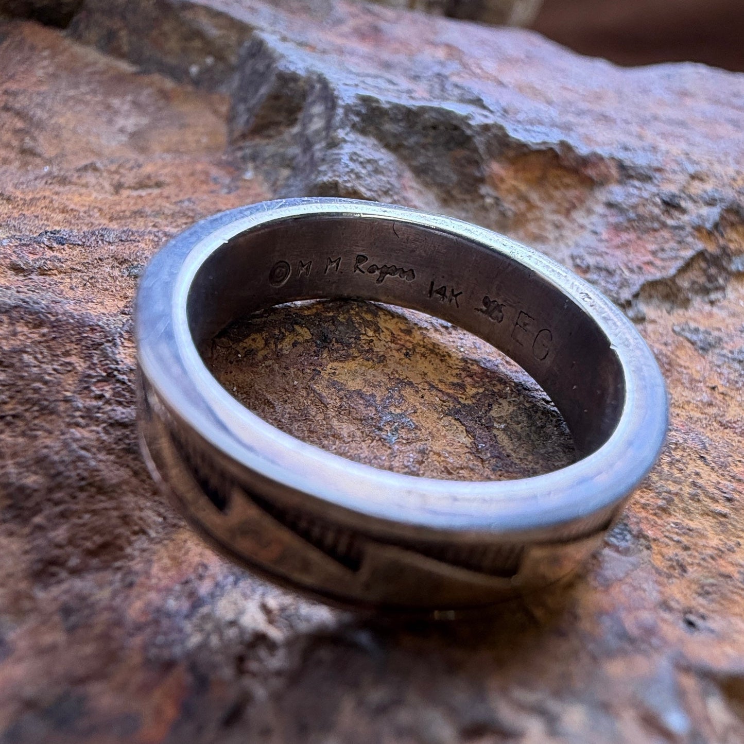 A silver-toned metal ring with a textured, ridged exterior and a stamped interior, resting on a textured brown and orange rock.