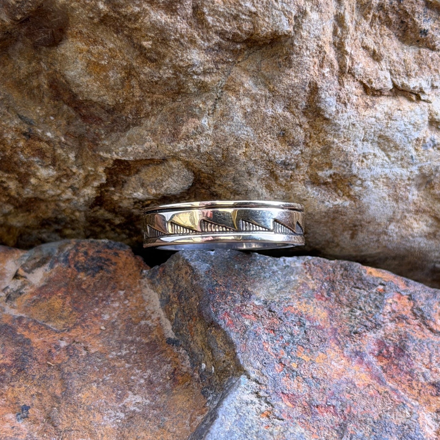 A silver band ring with a geometric chevron pattern sits nestled between textured orange and brown rocks.
