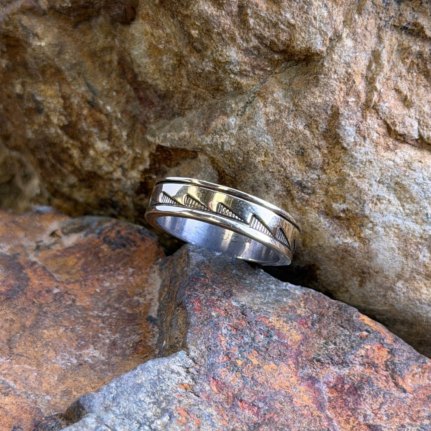A silver band ring with a textured geometric pattern, resting on rough, natural rocks outdoors.