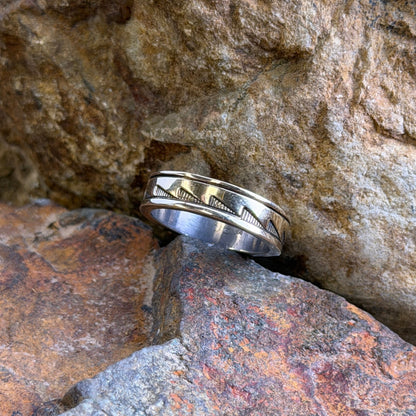 A silver band ring with a textured geometric pattern, resting on rough, natural rocks outdoors.