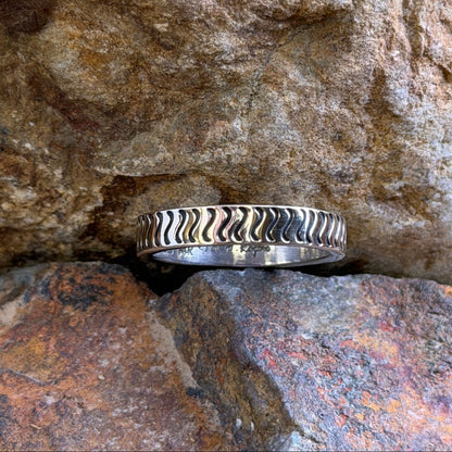 A silver ring with a textured, wavy chevron pattern sits nestled in a crevice between natural brown and orange rocks.