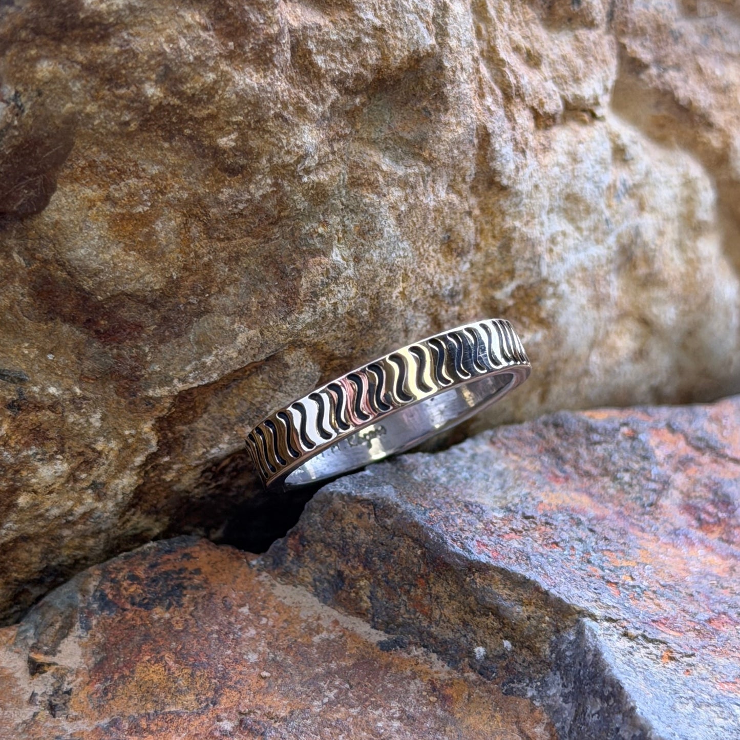 A silver ring with a wavy pattern resting on stacked natural rocks.