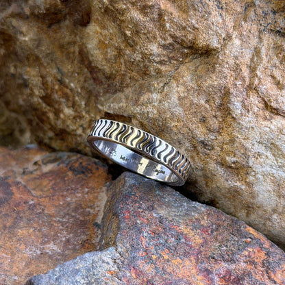 A silver ring with a wavy, textured pattern sits nestled between natural brown and orange rocks.