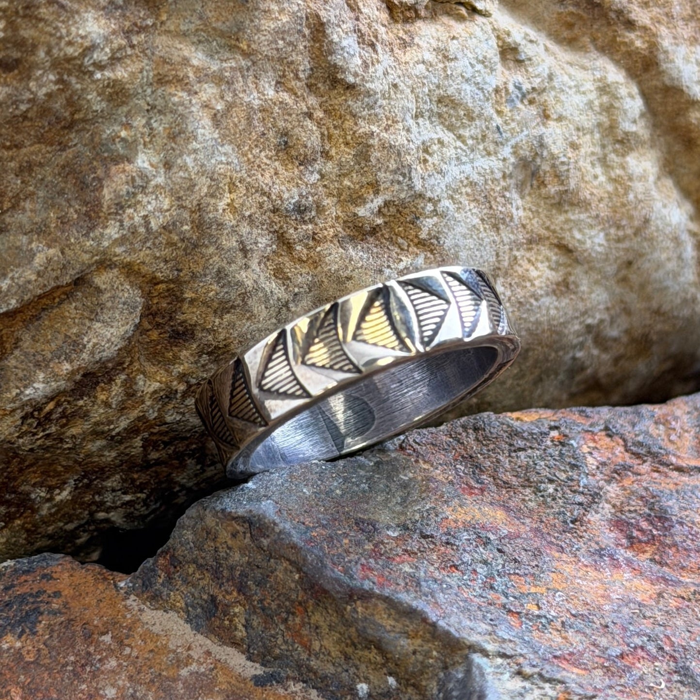 A silver ring with a geometric, faceted triangular pattern on its band, resting on textured natural rocks.