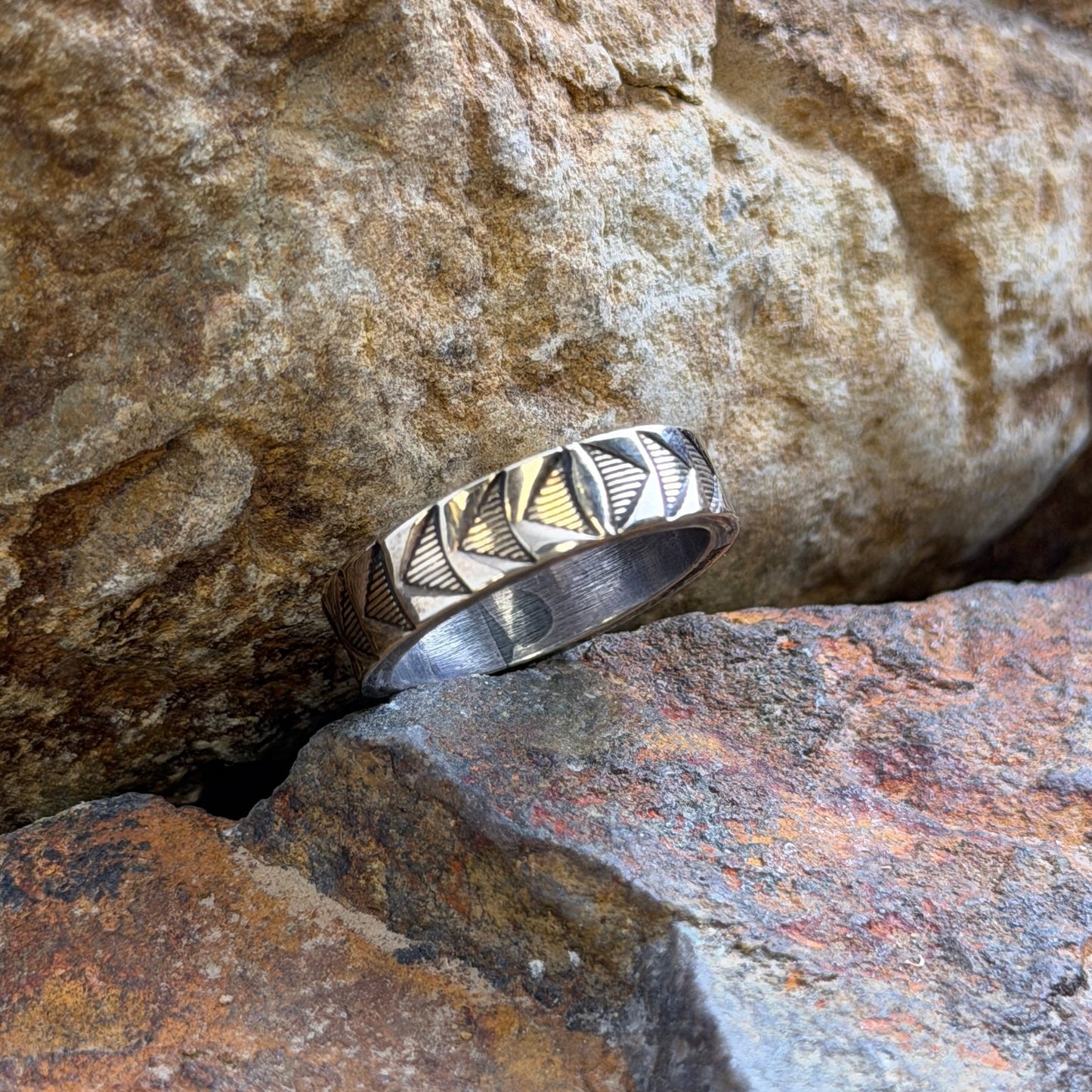 14k and sterling silver ring with a geometric, triangular arrow pattern on its band, resting on textured natural rocks. 