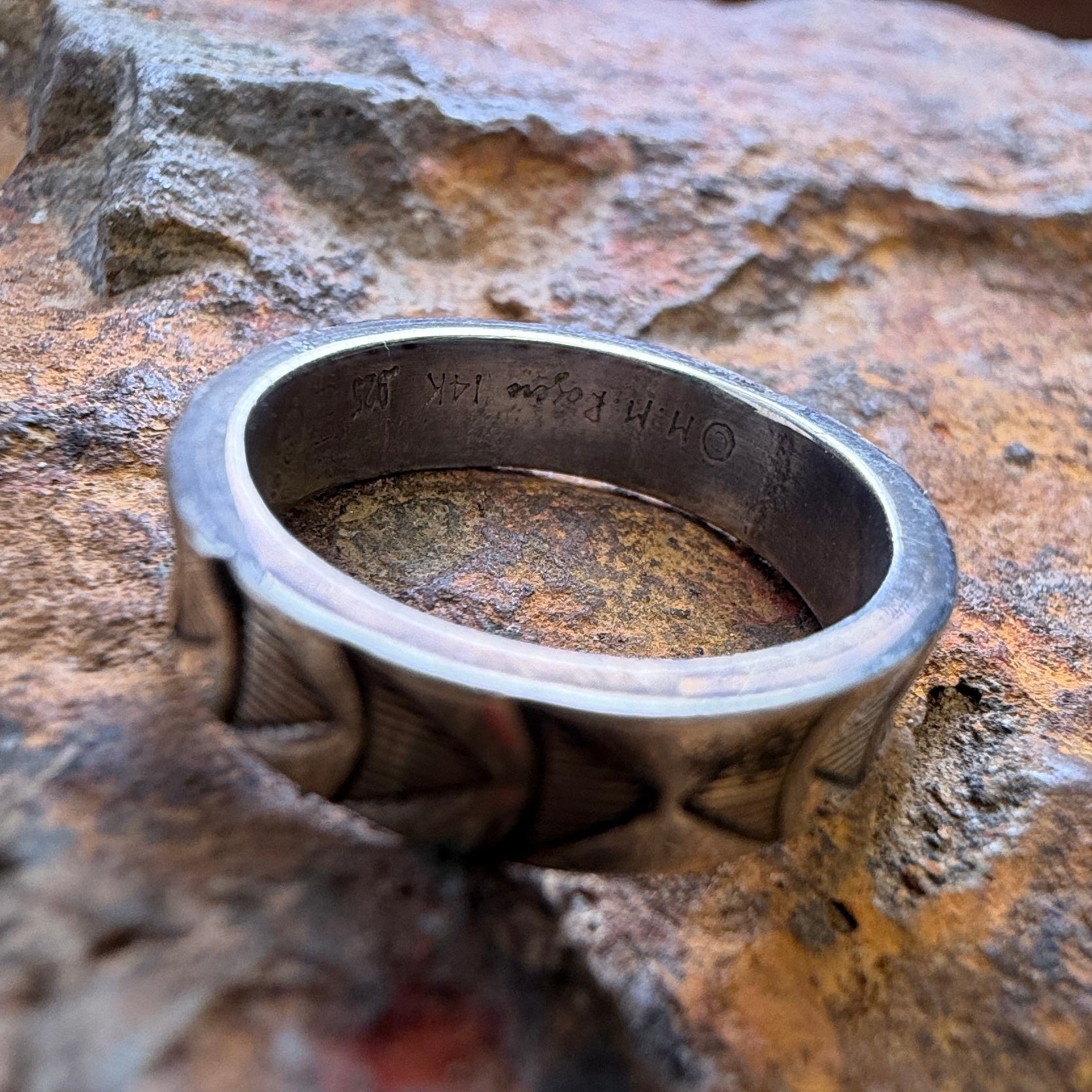 A silver-toned metal ring with a textured, geometric patterned band, stamping on the inside, resting on a textured brown and orange rock.