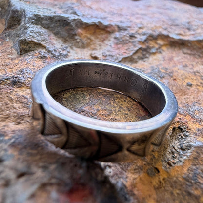 A silver-toned metal ring with a textured, geometric patterned band, stamping on the inside, resting on a textured brown and orange rock.