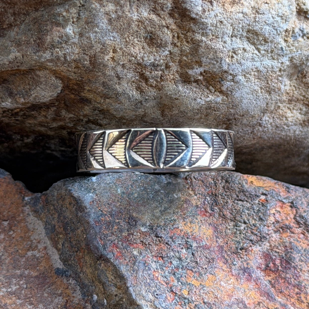 A silver geometric band ring with inlaid dark accents, resting on a textured orange-brown rock against a larger brown rock background.