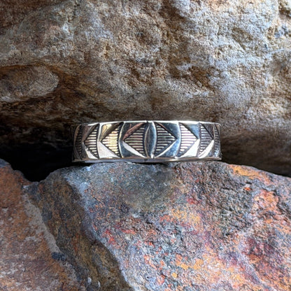 A silver geometric band ring with inlaid dark accents, resting on a textured orange-brown rock against a larger brown rock background.
