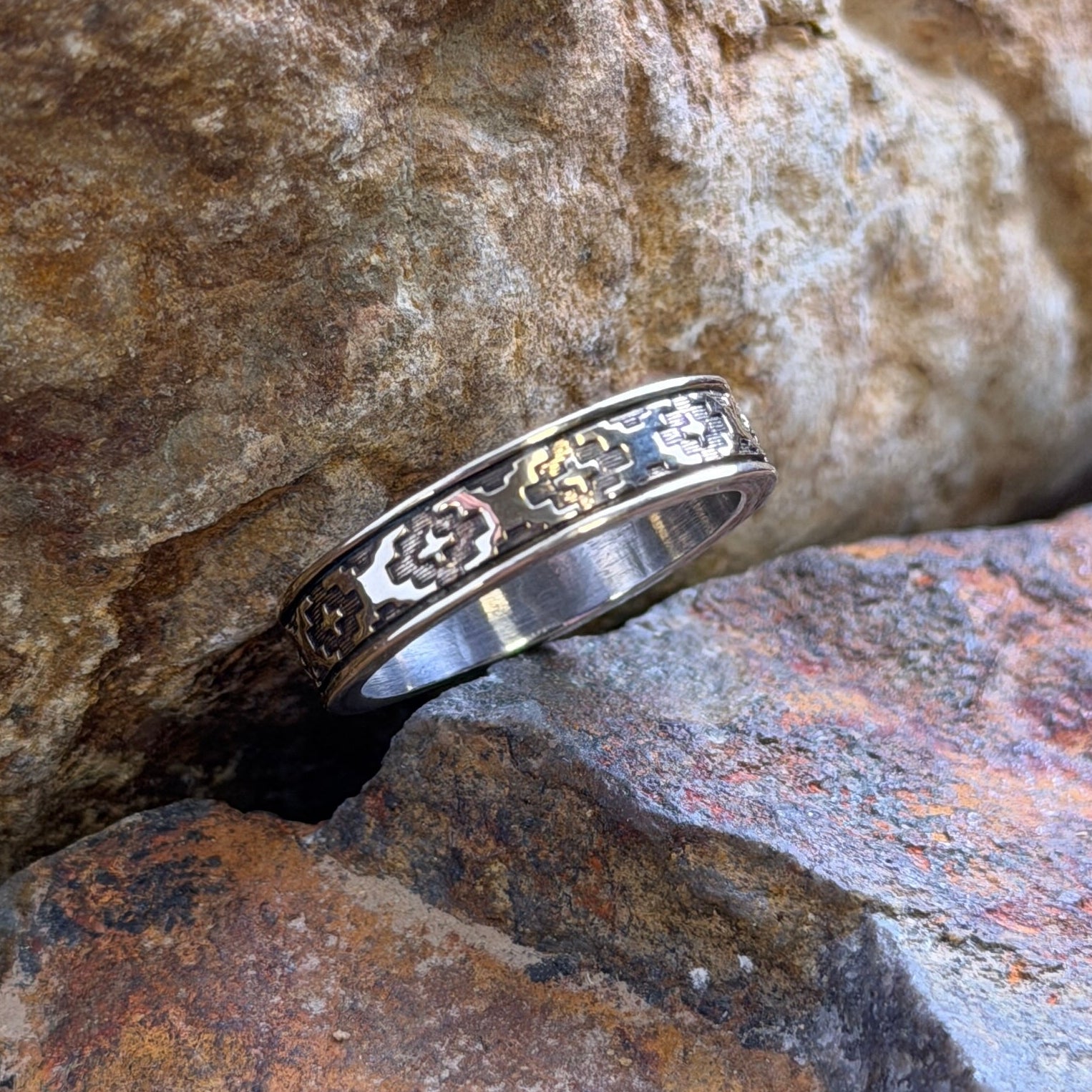 A silver & 14k gold band ring with a repeating southwest motif engraving, resting between textured brown and orange rocks.