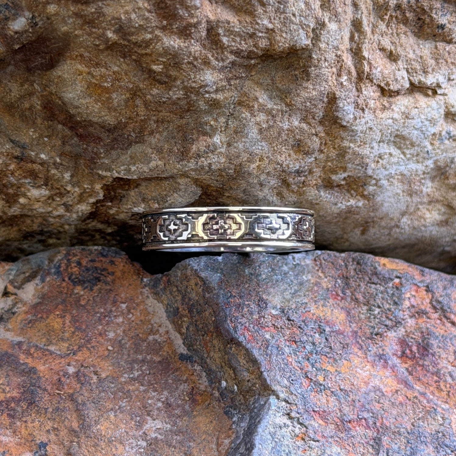 A silver-toned ring with a repeating geometric pattern of small gold-toned diamonds, resting on a textured brown and orange rock.
