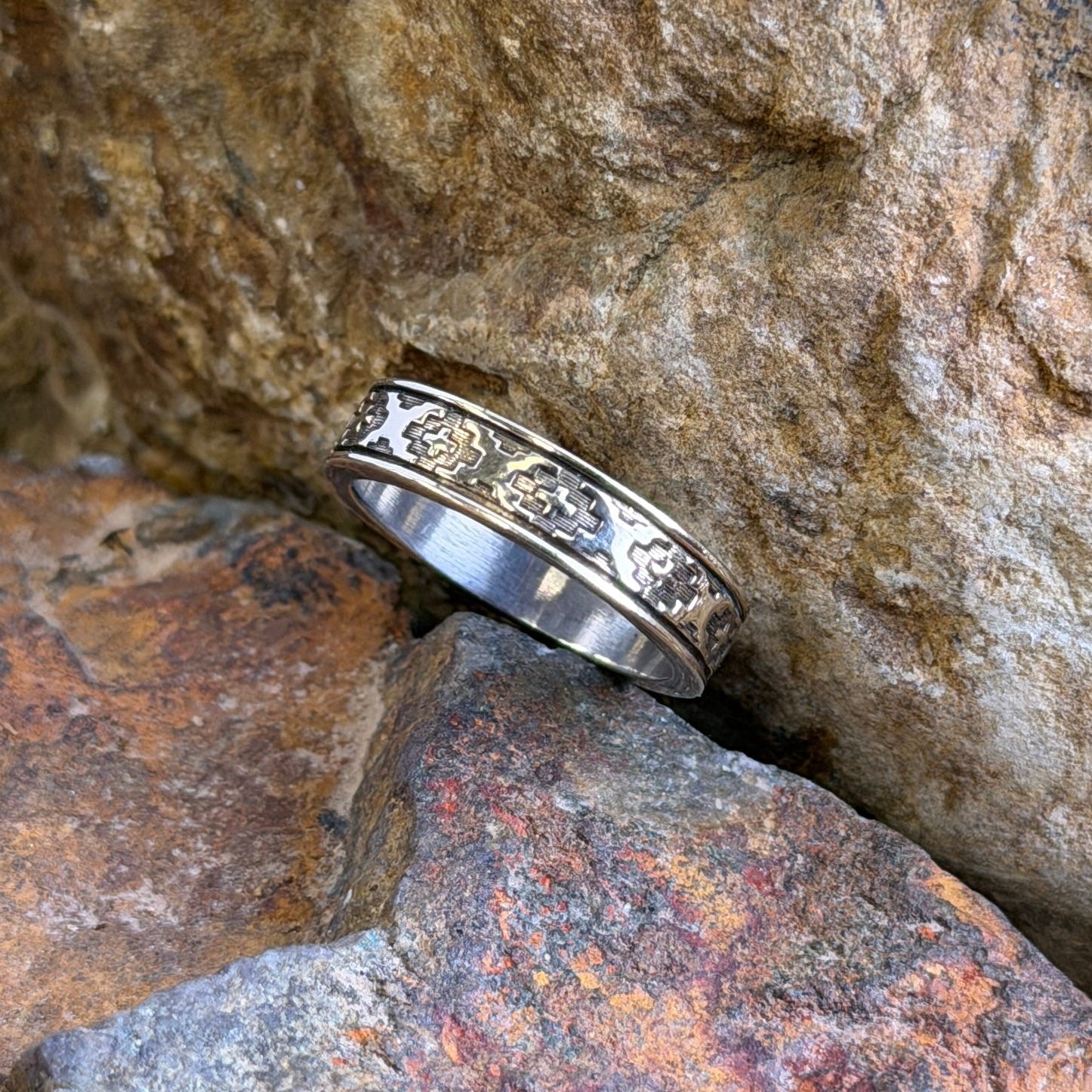 A silver band ring with a textured geometric pattern, resting on textured orange and brown rocks.