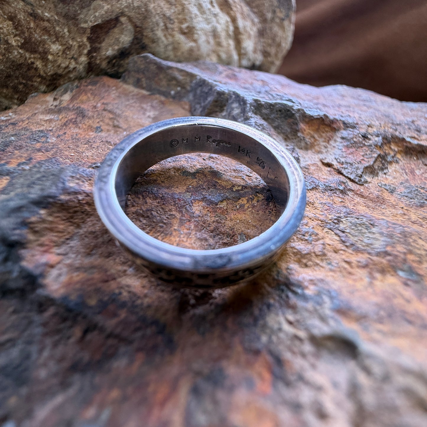 A silver-toned metal band ring with engraved text inside, resting on a textured brown and orange rock surface.