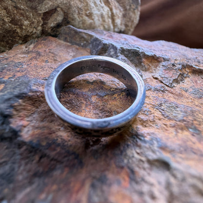 A silver-toned metal band ring with engraved text inside, resting on a textured brown and orange rock surface.