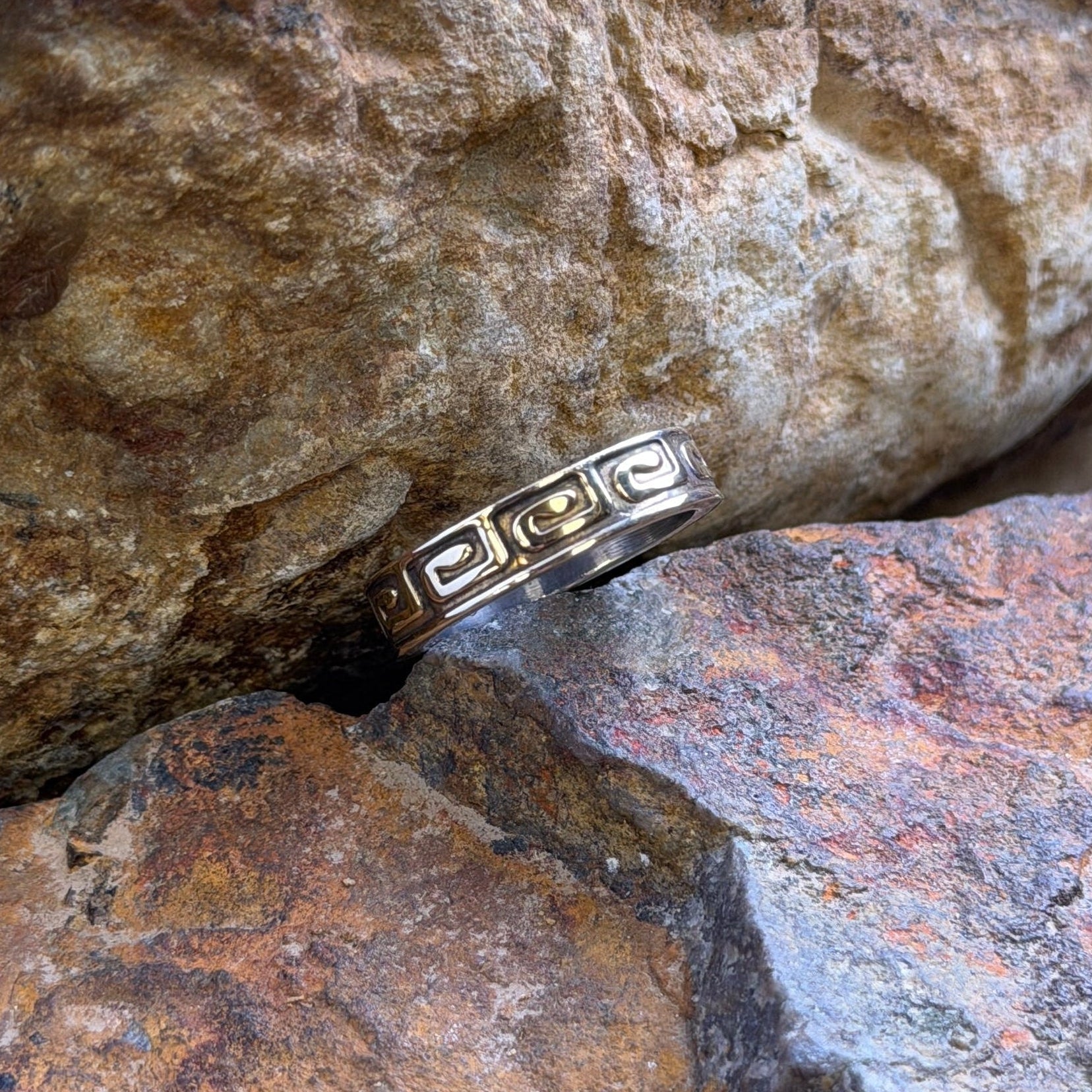 A silver-toned ring with a Greek key pattern and small clear stones, resting between two textured natural rocks.