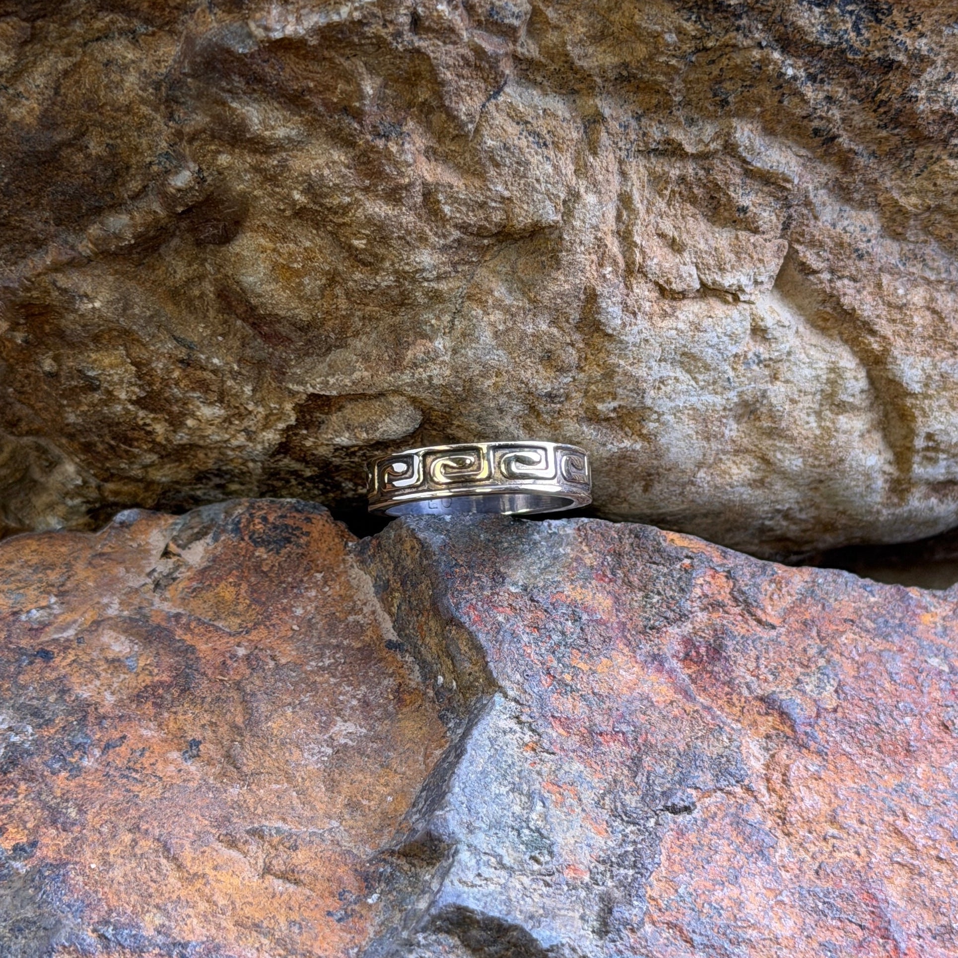 A silver band ring with engraved Greek key patterns, resting on a textured brown and orange rock.